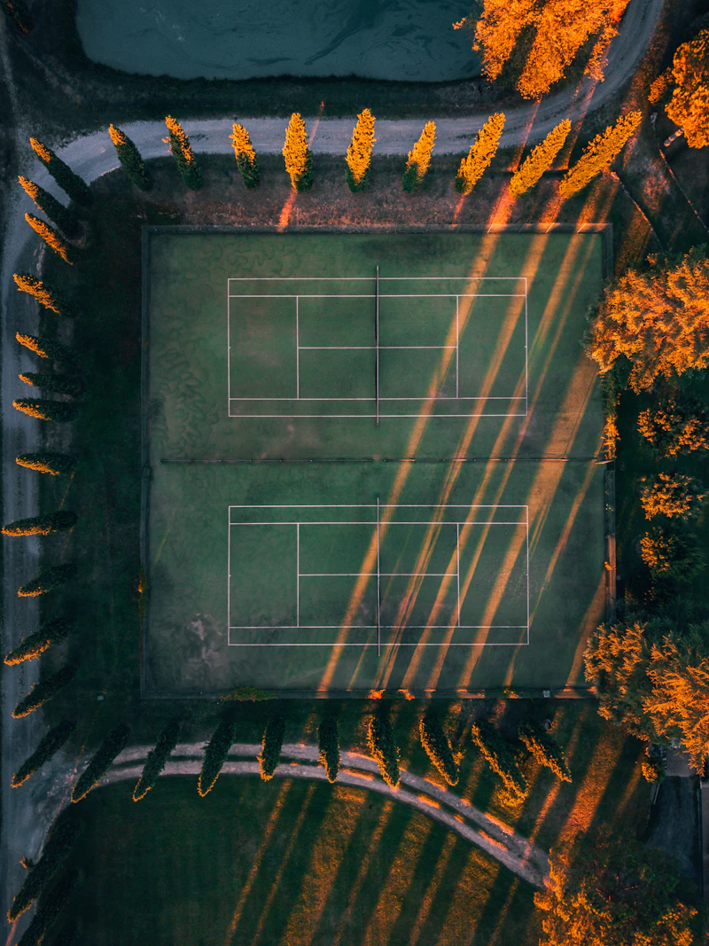 Two tennis courts seen from above, with trees and long shadows at sunset.